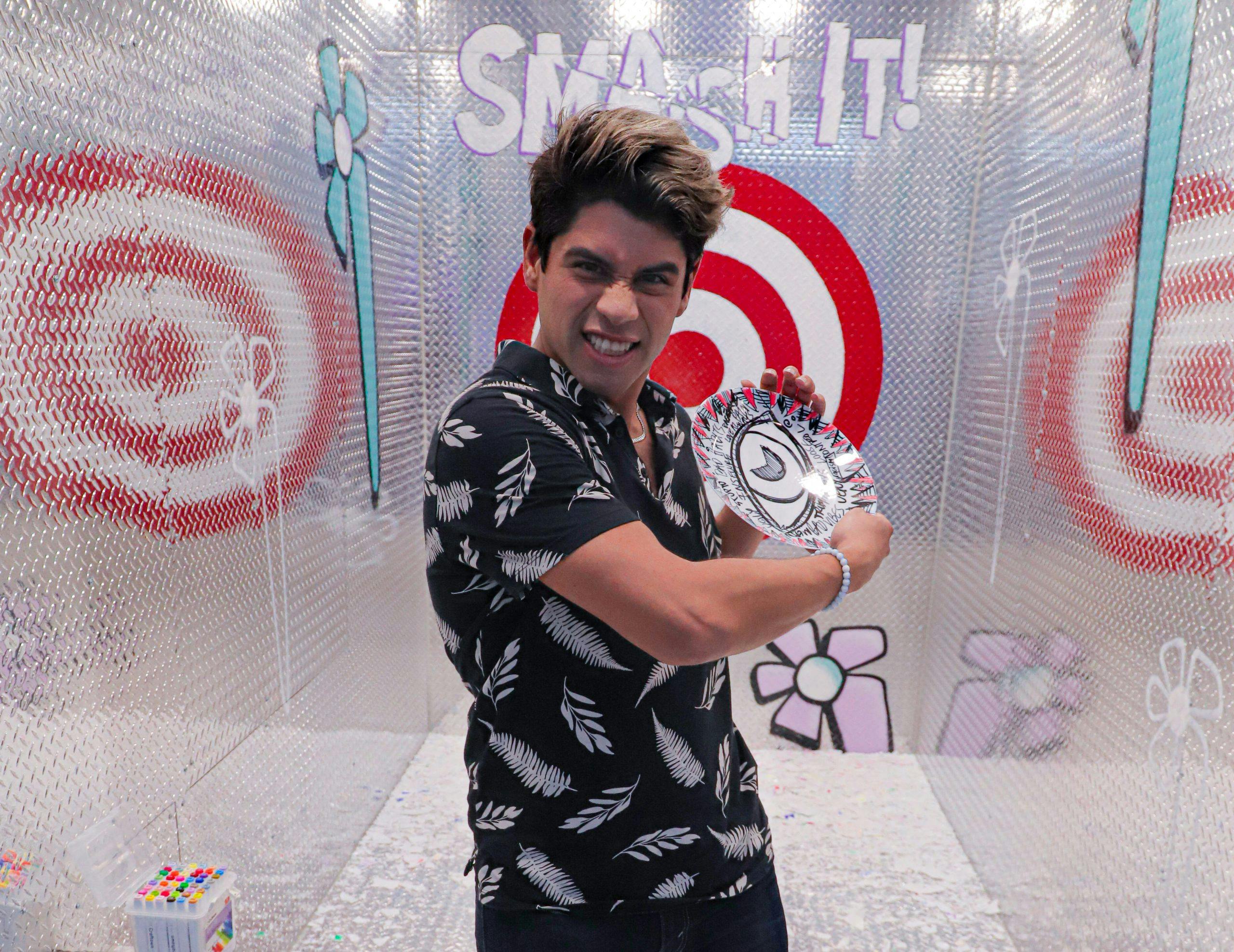 Persona con camicia a fantasia che fa un'espressione feroce mentre tiene in mano un piatto in una stanza con testo "SMASH IT!" e disegni di bersagli.