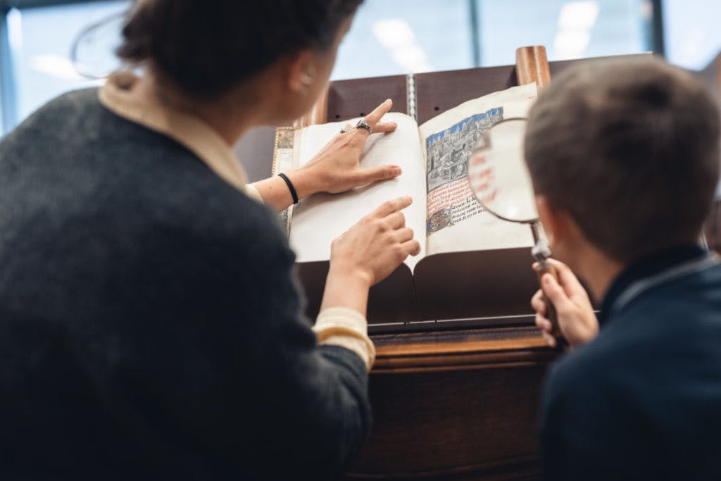 Un adult assenyala un llibre quan un nen utilitza una lupa per examinar la pàgina en un entorn de biblioteca.