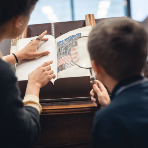 En vuxen pekar på en bok som barn och använder ett förstoringsglas för att undersöka sidan i en biblioteksinställning.