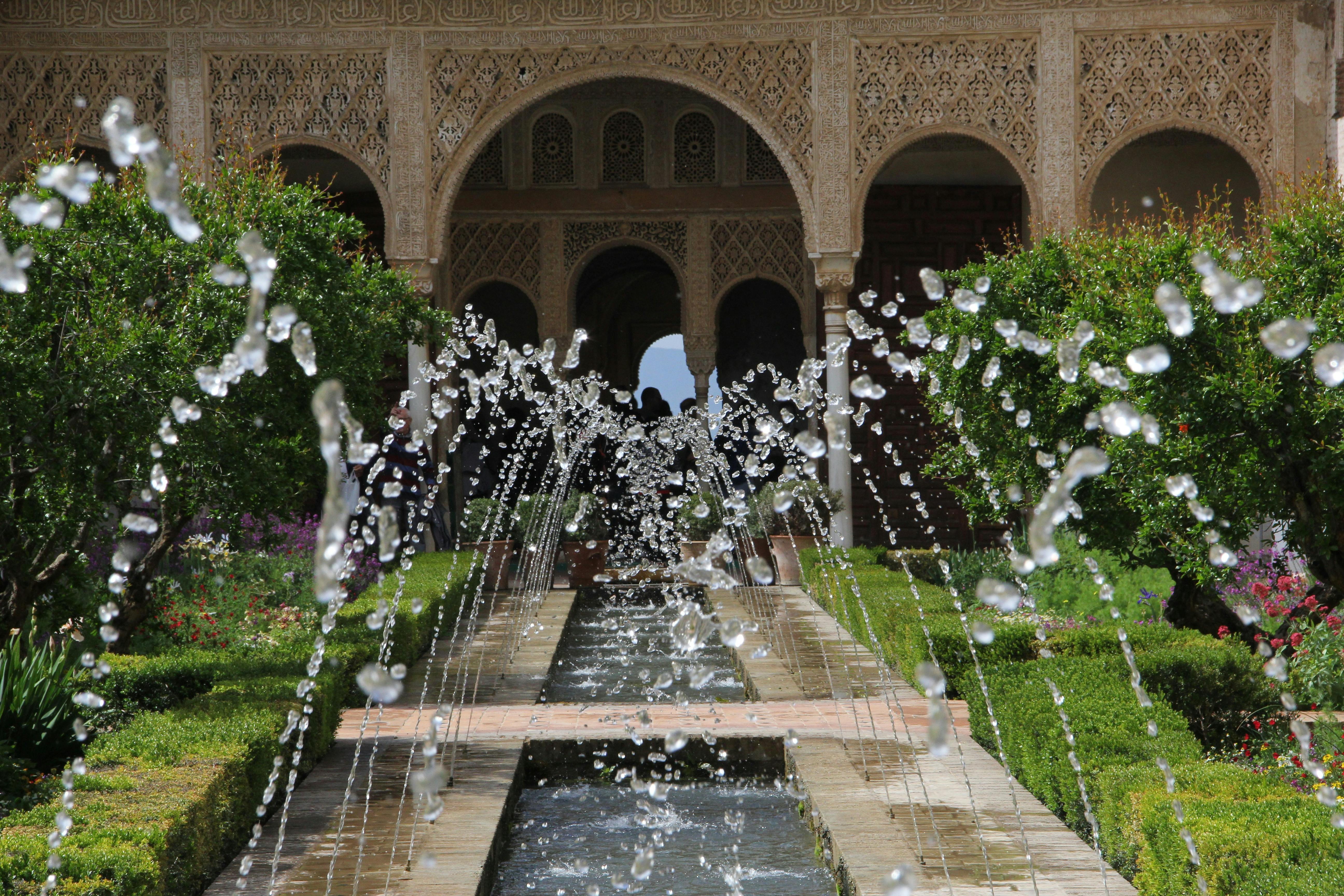 Ornate garden with arched building in the background; central pathway features a fountain with jets of water spraying upward.