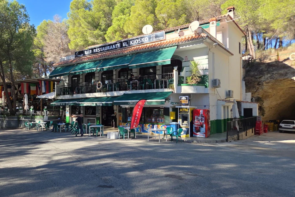 An exterior view of a restaurant called "Bar Restaurante El Kiosko", featuring green awnings, outdoor seating and surrounding trees.