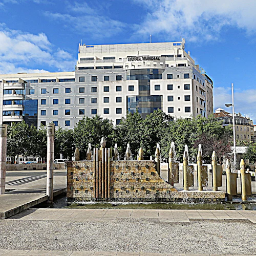 Modern byggnad med många fönster bakom ett trädkantat område med en fontän med vertikala cylindriska strukturer. Blå himmel ovanför.
