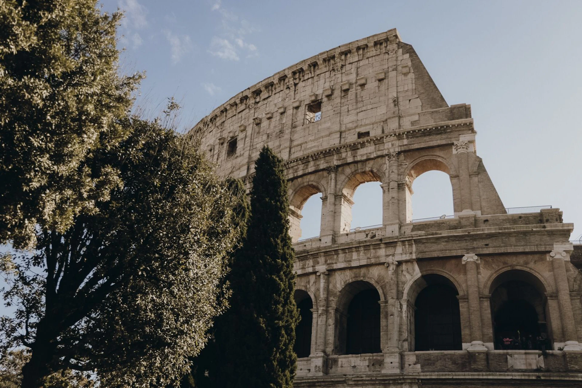 The Colosseum in Rome, partially obscured by trees, under a clear blue sky.