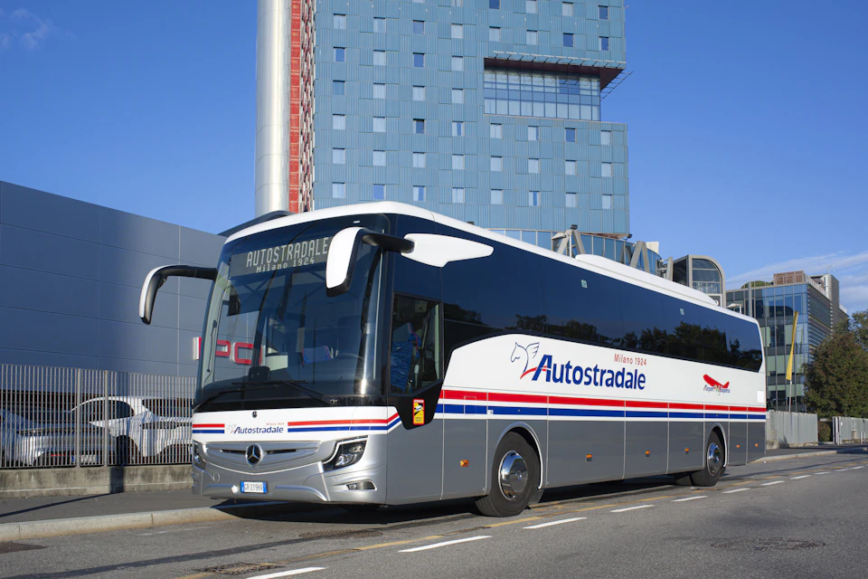 A silver Autostradale bus is parked in front of a modern, multi-story building with a blue and red facade under a clear sky.