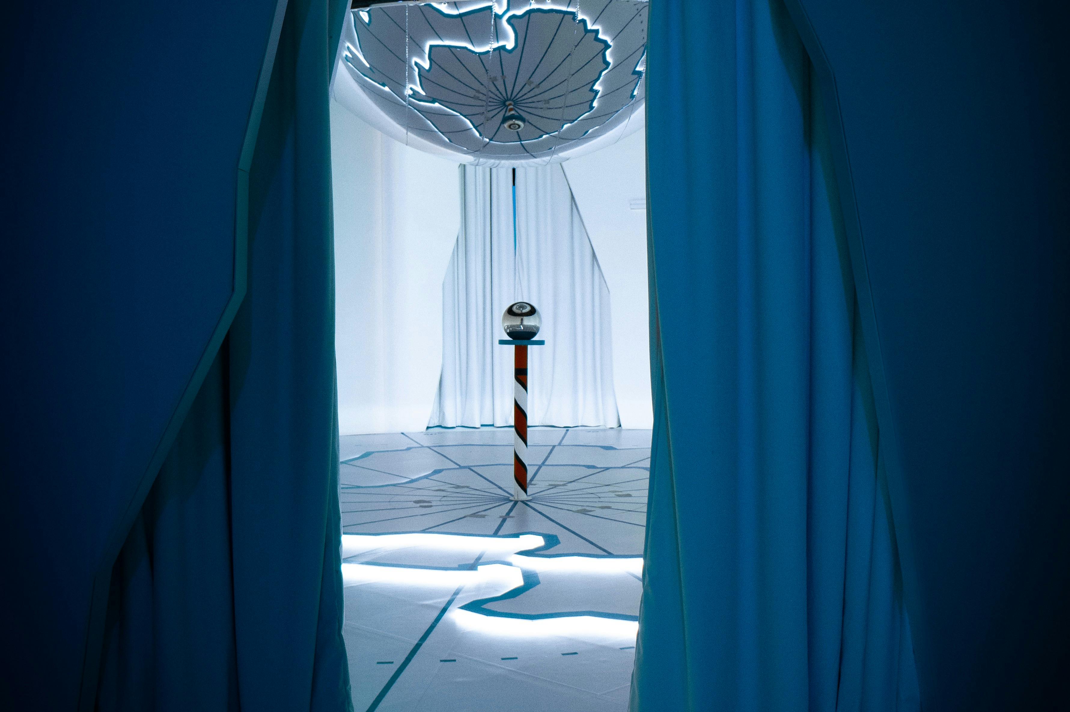 Futuristický pokoj s modrými závěsy, vzorovanou podlahou, designem zavěšeného stropu a středovou tyčí s koulí připomínající oko nahoře.