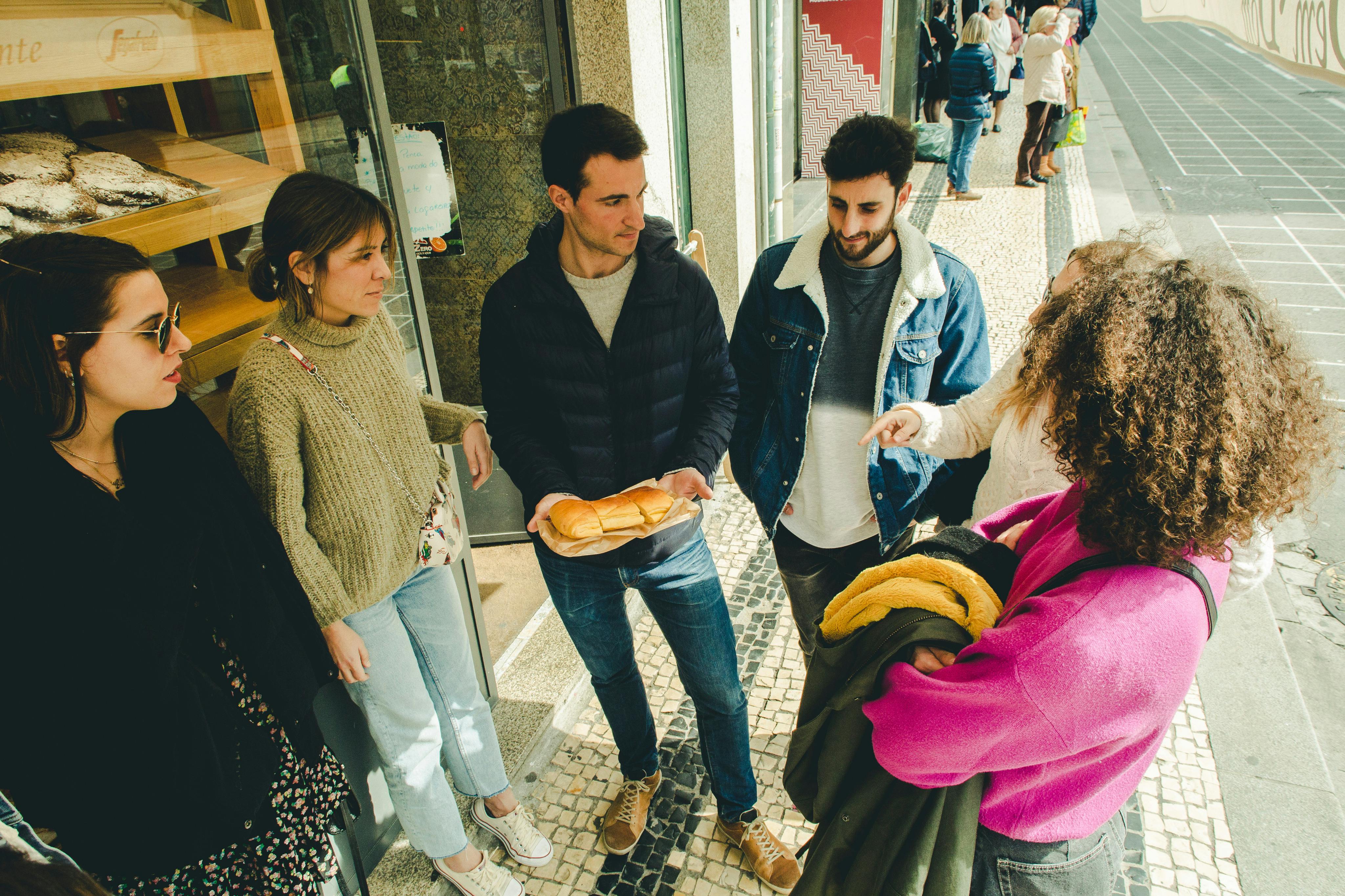 Een groep van vijf mensen staat voor een winkel, één persoon houdt een stokbrood vast. Eén persoon wijst naar een andere persoon.