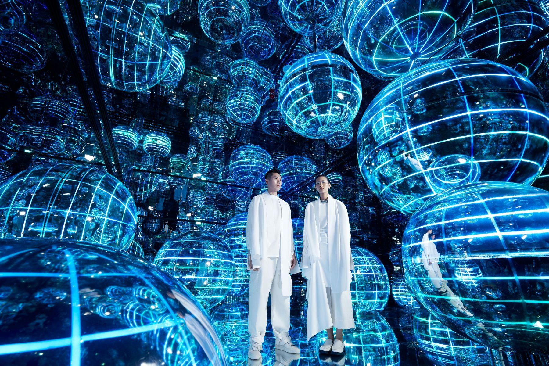 흰색 옷을 입은 두 사람이 반짝이는 파란색 구형 구조물과 거울로 가득 찬 방 한가운데 서 있습니다.