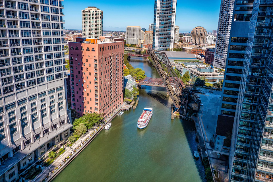 A river winding through a city with a white and red boat, surrounded by tall buildings, and a raised drawbridge.