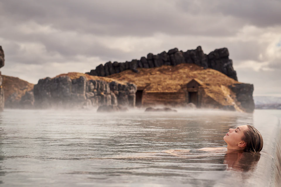 Person floating in water with rocky hills and old stone buildings in the background under a cloudy sky.