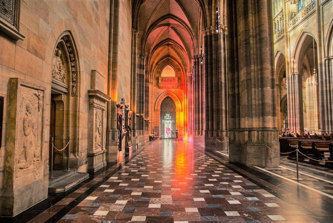 Gothic-style cathedral hallway with high arches, checkerboard floor, and warm lighting illuminating an entrance in the distance.