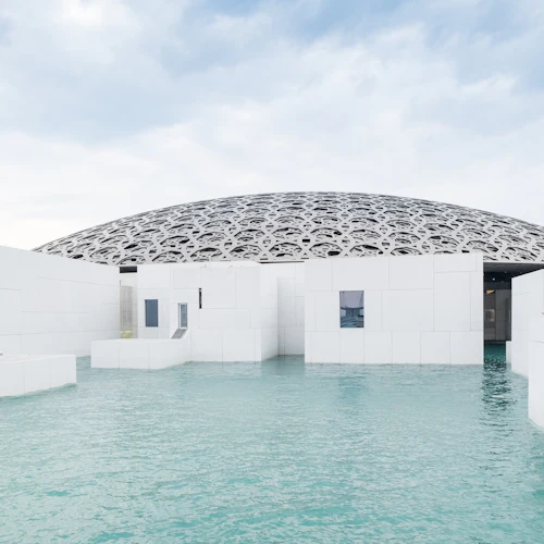 Weiße geometrische Gebäude, umgeben von Wasser, mit einer verschlungenen Kuppelstruktur im Hintergrund und einem blauen Himmel.