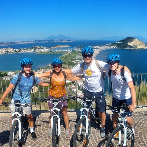 Four people wearing helmets stand with bicycles on a scenic overlook with coastal views and distant islands in the background.
