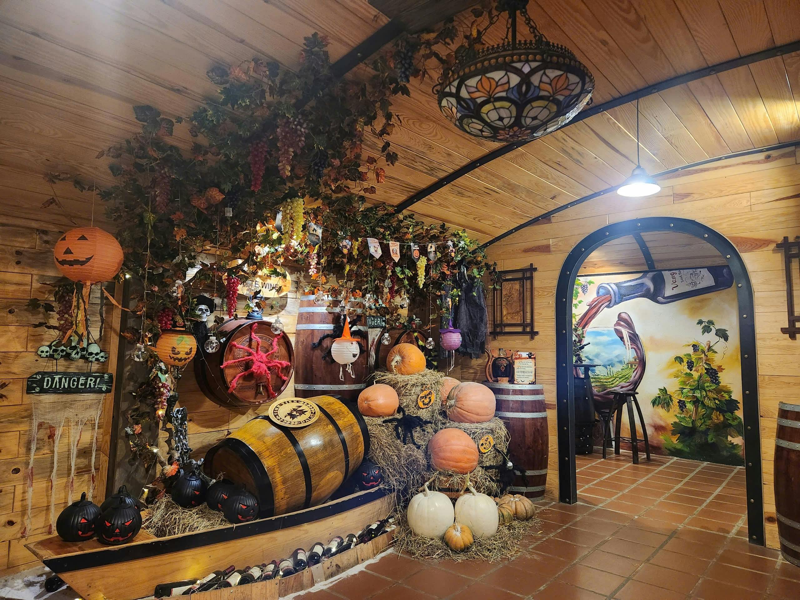 A cozy, decorated room with wooden barrels, pumpkins, hanging vines, and festive ornaments under a wooden ceiling.