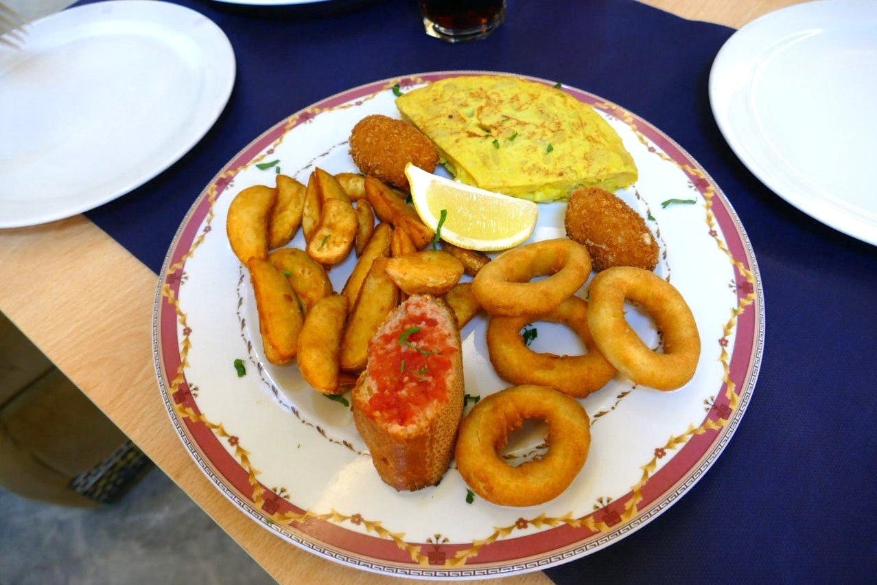 Une assiette de tapas assorties, comprenant des quartiers de pommes de terre, des rondelles d'oignon, des croquettes, des tortillas et du pain à la tomate, garnie d'un quartier de citron.