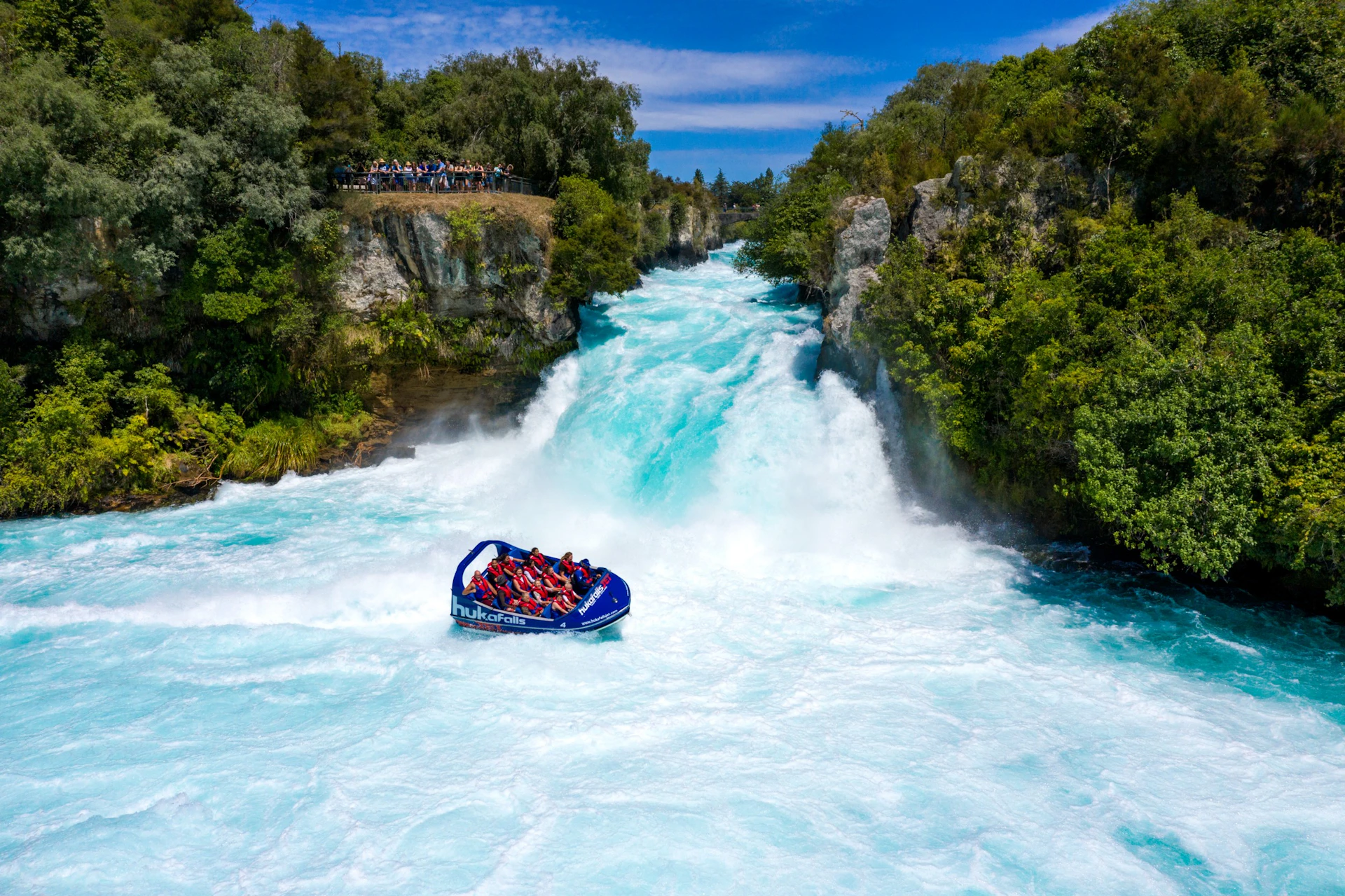 A jet boat navigates a fast-flowing river near a waterfall surrounded by greenery and observed by a distant group of people.