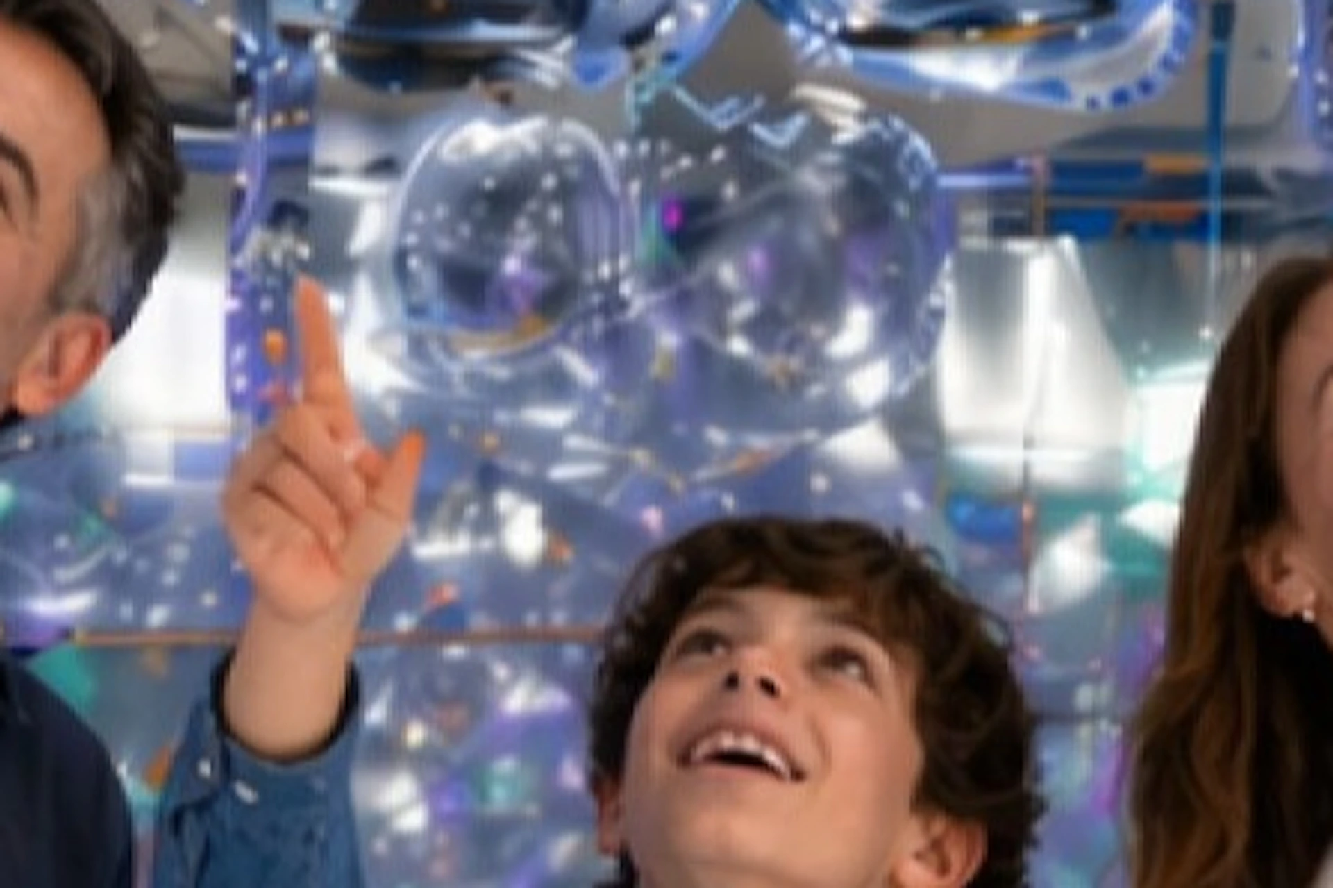 Three people, two adults and a child, smiling and pointing at reflective silver balloons hanging above them indoors.