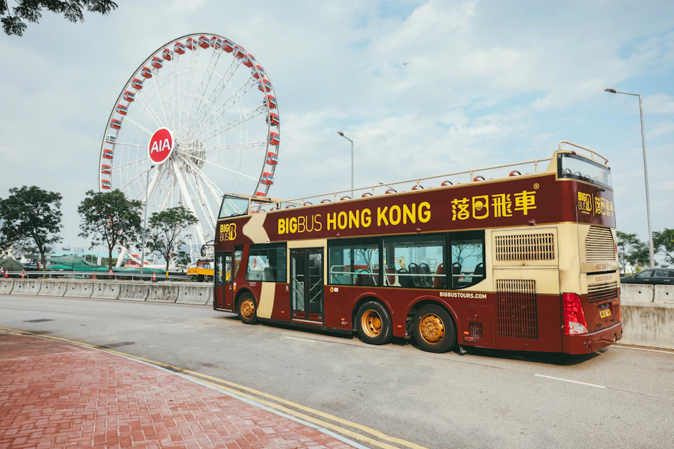 A red and beige double-decker bus labeled "Big Bus Hong Kong" is parked near a large Ferris wheel in a city area.