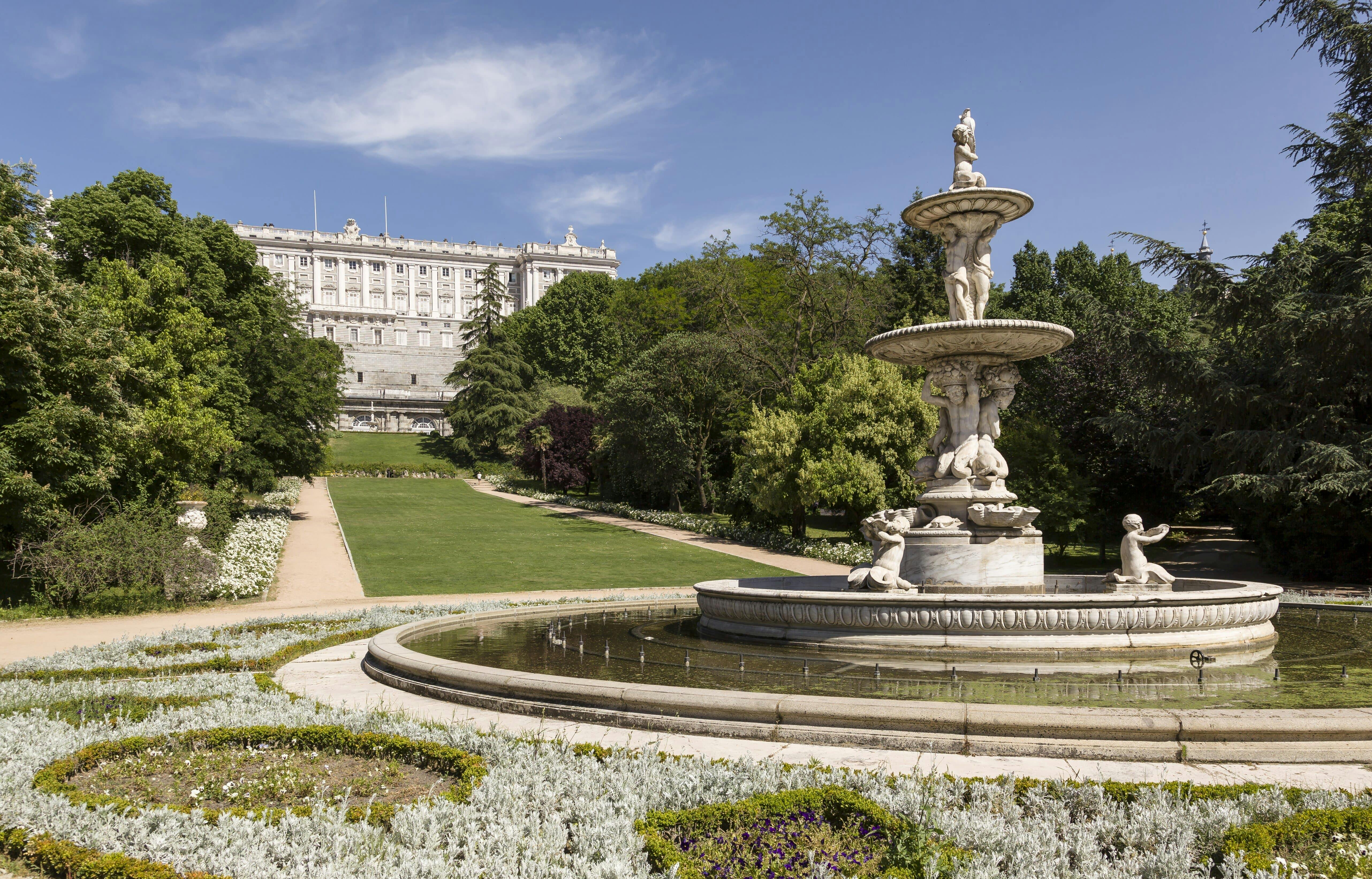 Za svěží zahradou a velkou kamennou fontánou za slunečného dne stojí historický palác s modrou oblohou nad hlavou.