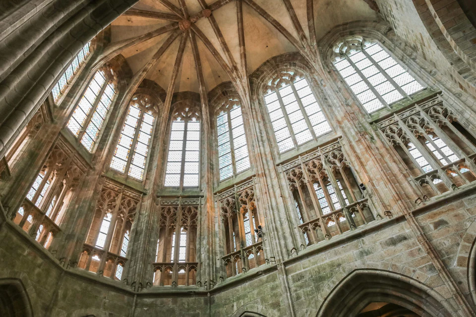Interior of a Gothic cathedral with tall, arched stained glass windows and ornate stone detailing under a vaulted ceiling.