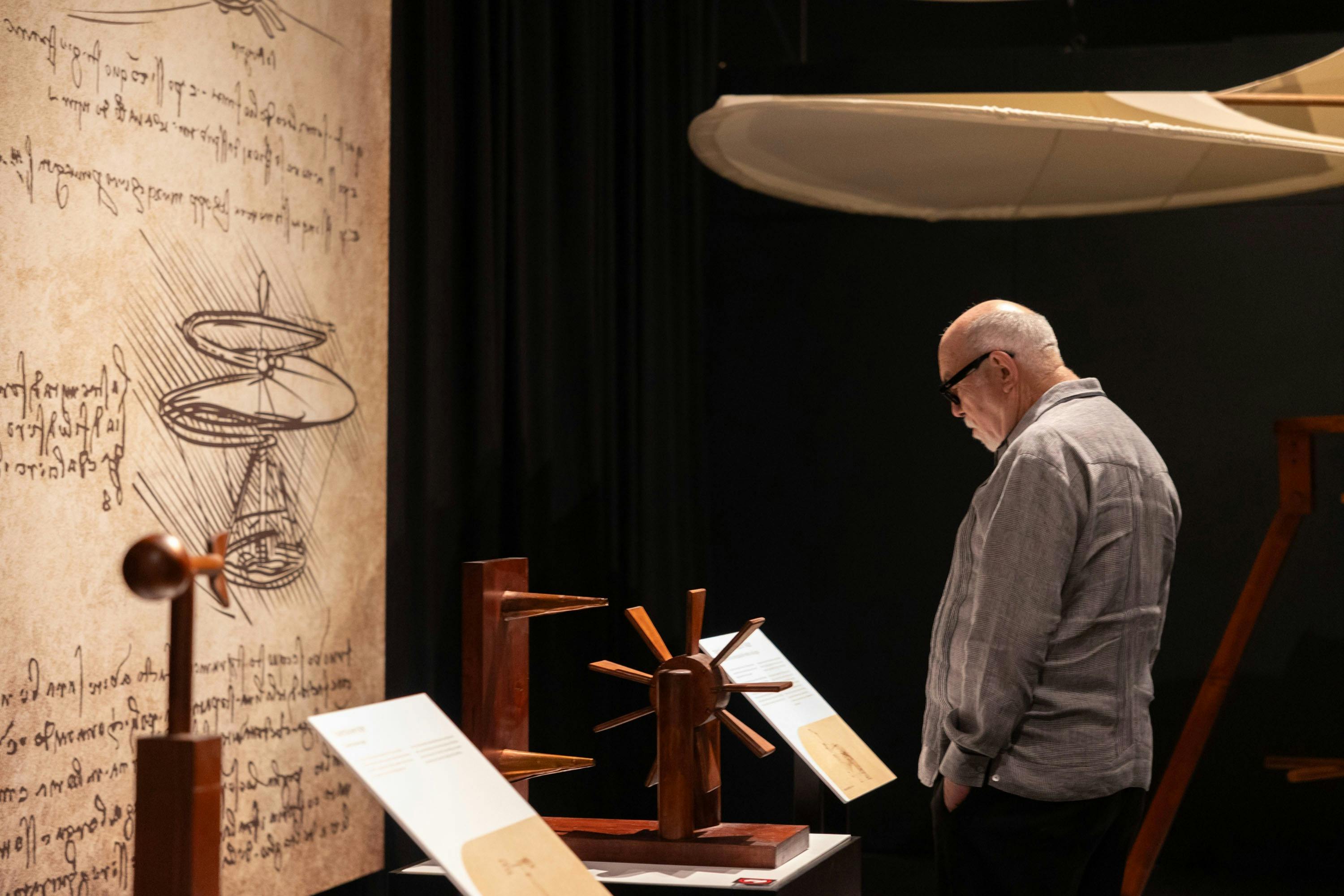 En mand i en grå skjorte ser på træmodeller, der vises i en svagt oplyst udstilling. En stor struktur hænger fra loftet.