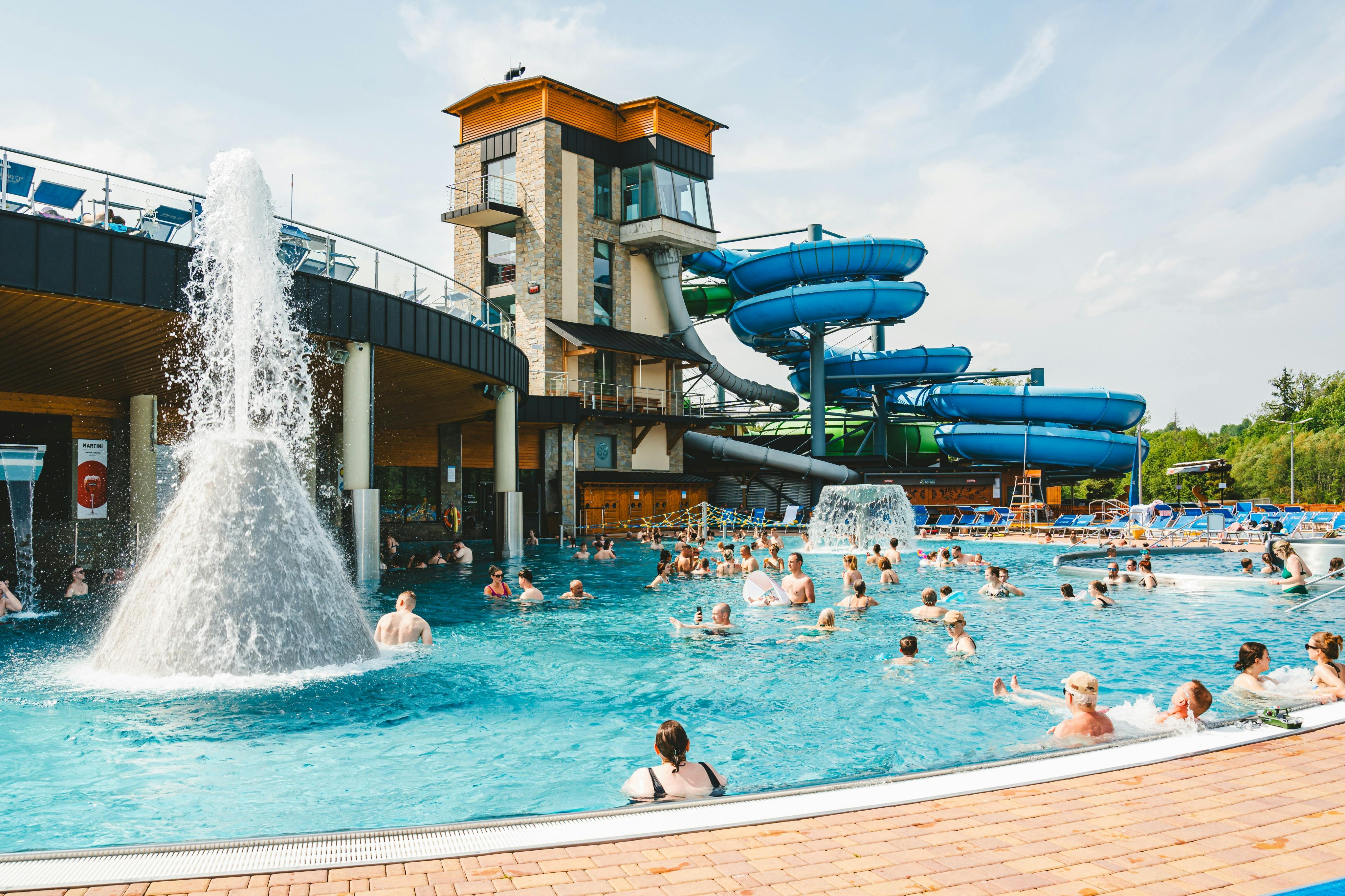 ウォータースライダー、噴水、晴れた空の下の複数階建ての建物がある混雑したプールエリア。