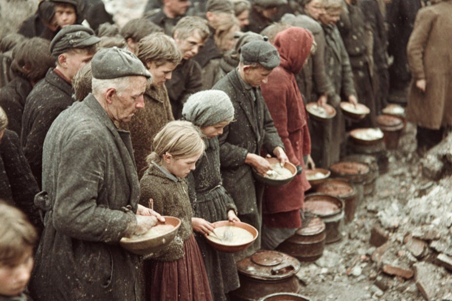 Eine Gruppe von Menschen, die in einer Reihe stehen und Schüsseln in der Hand halten, in abgenutzter Kleidung, in einer Umgebung, die wie ein Außenbereich aussieht.