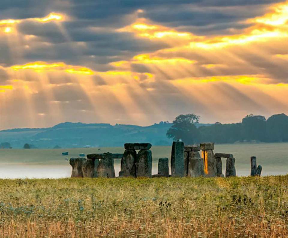 Stonehenge at sunrise with sunbeams piercing through the clouds, illuminating the ancient stones and the grassy landscape.