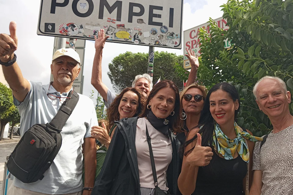 A group of six people smiling and posing in front of a sign that reads 'Pompeii,' with trees and greenery in the background.