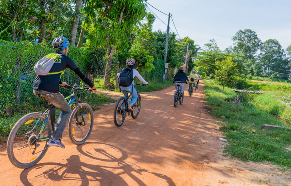 Four people riding bicycles on a dirt road surrounded by greenery and trees under a clear sky.