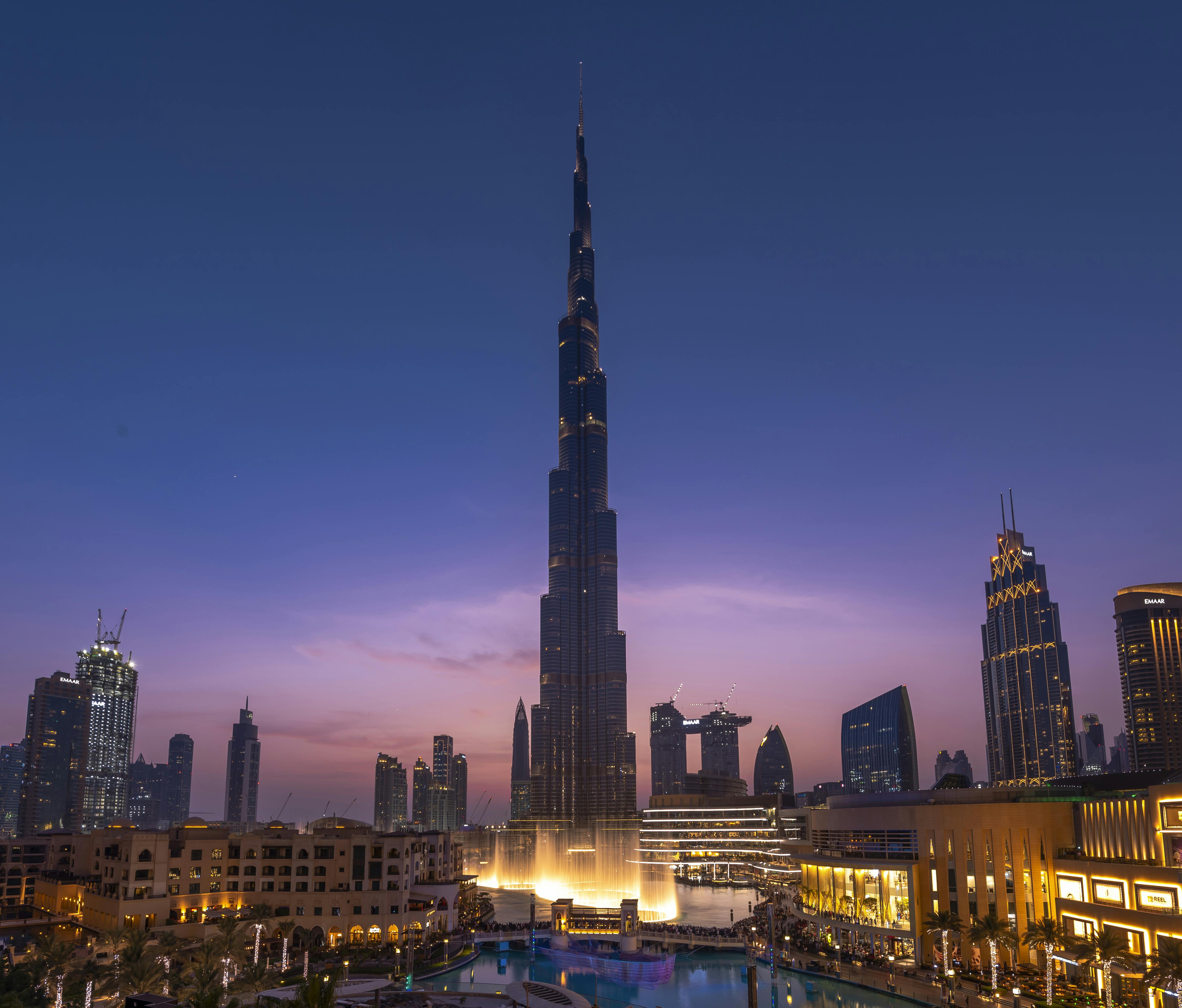 Vysoký osvětlený mrakodrap stojí za soumraku uprostřed panoramatu města s okolními budovami a odrazivým bazénem v popředí.