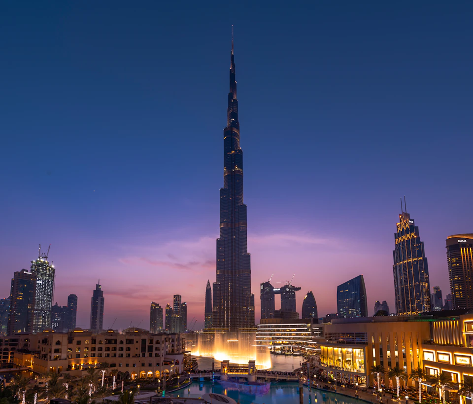 A tall, illuminated skyscraper stands amidst a cityscape at dusk with surrounding buildings and a reflecting pool in the foreground.