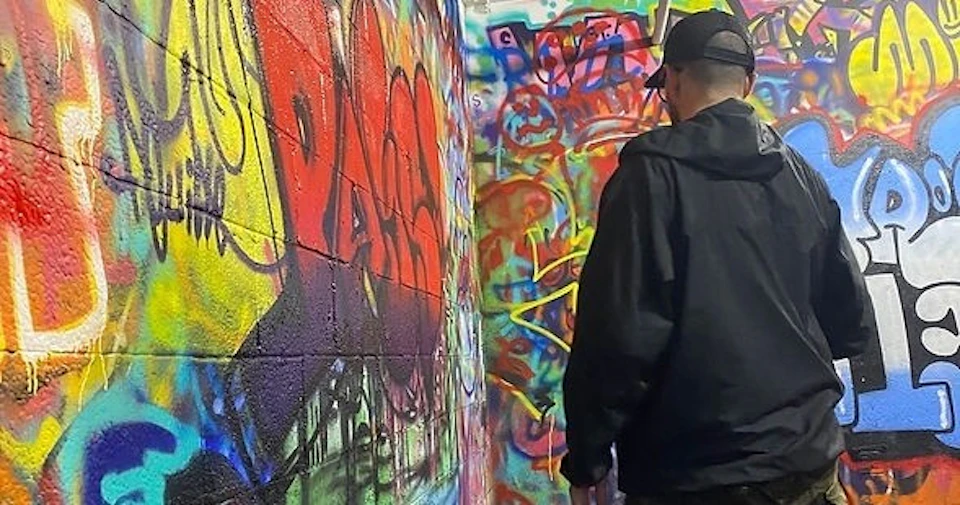 A person wearing a black jacket and cap standing in front of a wall covered with colorful graffiti.