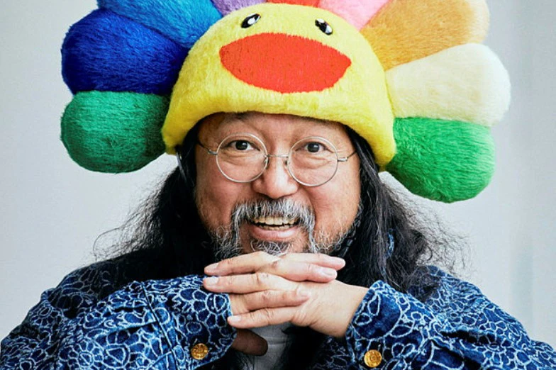 Person with round glasses, long hair, and a colorful plush hat shaped like a sunflower, smiling and resting hands under chin.