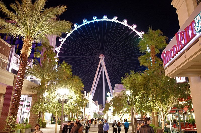 Le persone camminano di notte lungo una strada illuminata e costeggiata da palme, con una grande ruota panoramica illuminata sullo sfondo.