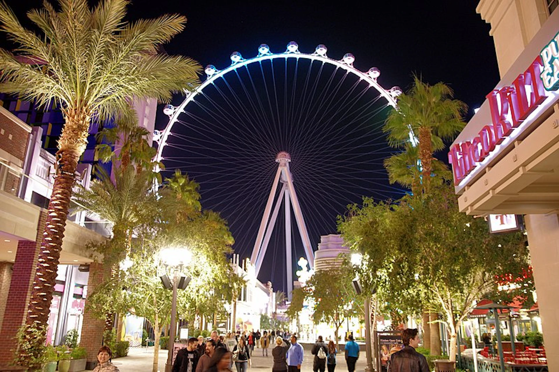 People walk along a brightly lit, palm tree-lined street at night with a large illuminated Ferris wheel in the background.