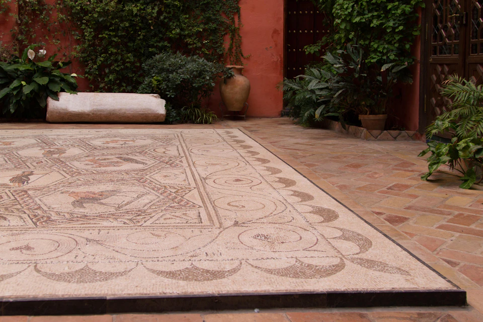 A courtyard with a detailed mosaic floor, surrounded by lush greenery, potted plants, and a red wall with an urn.