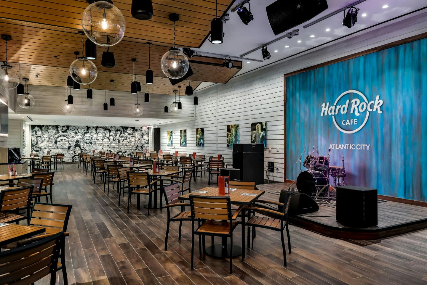 A modern restaurant with wooden tables and chairs, a stage with musical instruments, and a blue wall with "Hard Rock Café" signage.