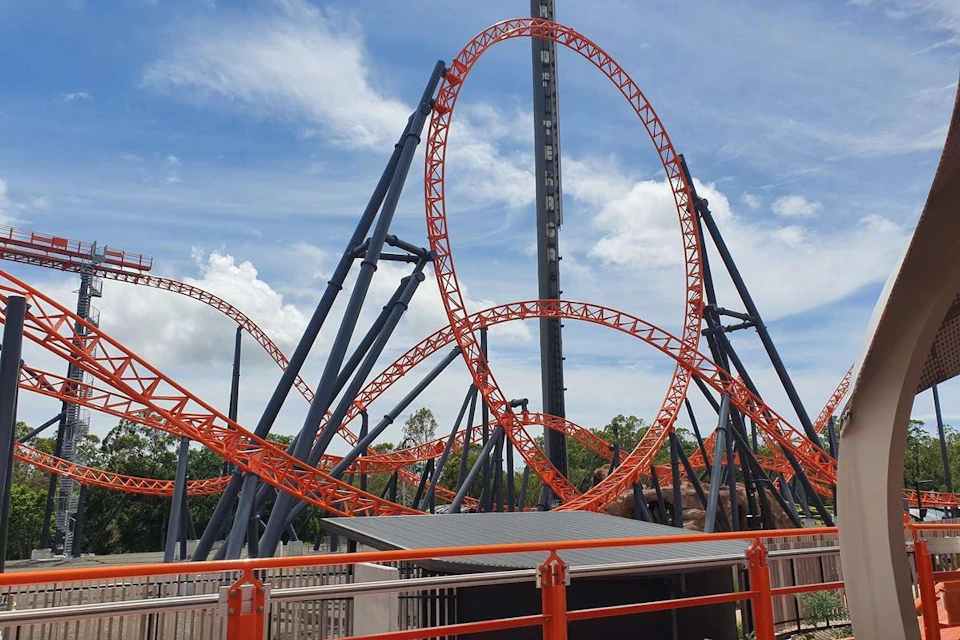 A roller coaster with orange tracks and loops against a blue sky with scattered clouds, surrounded by greenery and fencing.