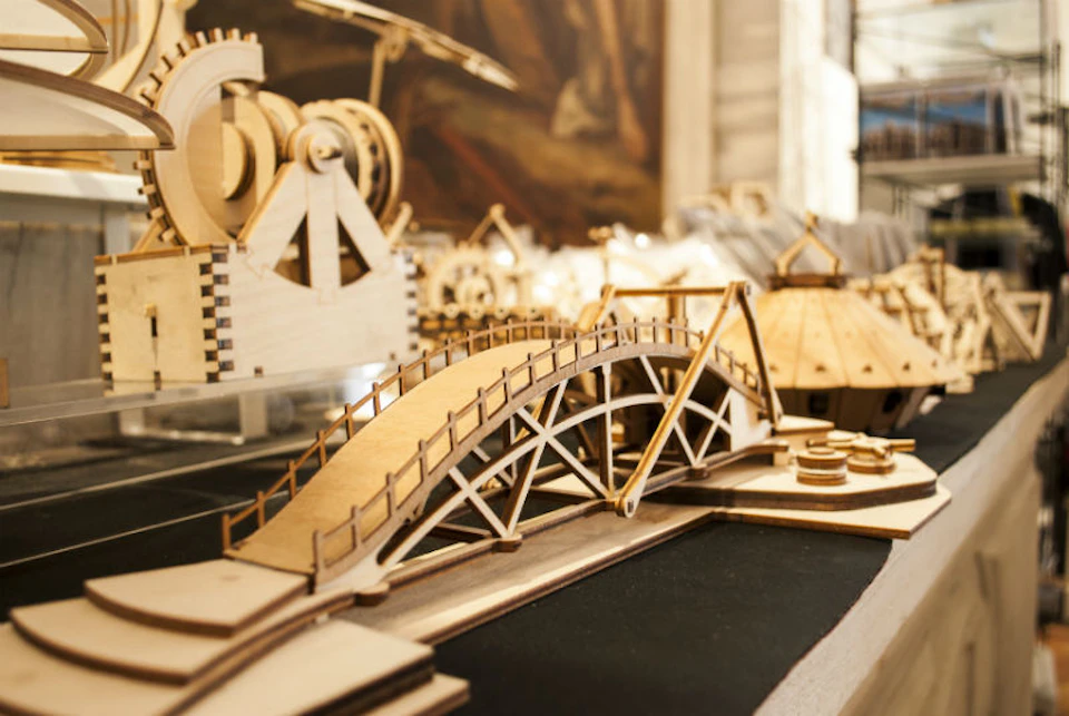 A detailed, wooden model of a bridge and machinery displayed on a table, with other wooden constructions in the background.