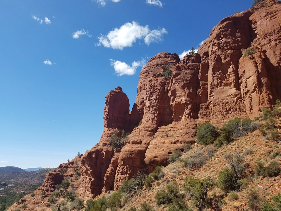 Rugged red rock formations under a blue sky with scattered clouds, surrounded by sparse greenery and desert vegetation.