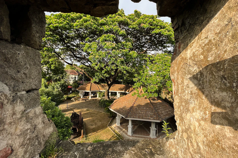 A view through a stone opening of a garden with gazebos and large trees, under a sunny sky, with a historical ambiance.