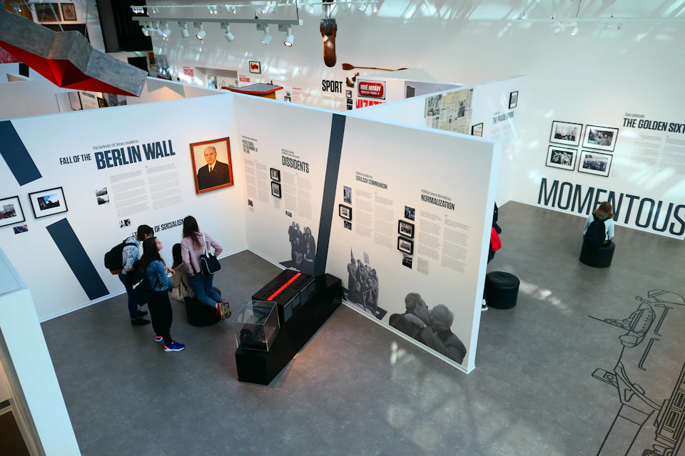 Visitors in a museum viewing exhibits about the Berlin Wall, dissidents, and normalization, with informational panels and photos.