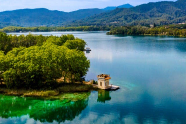 Un petit edifici groc situat en un moll, al costat d'un llac clar i tranquil, envoltat d'una frondosa vegetació i de muntanyes llunyanes.