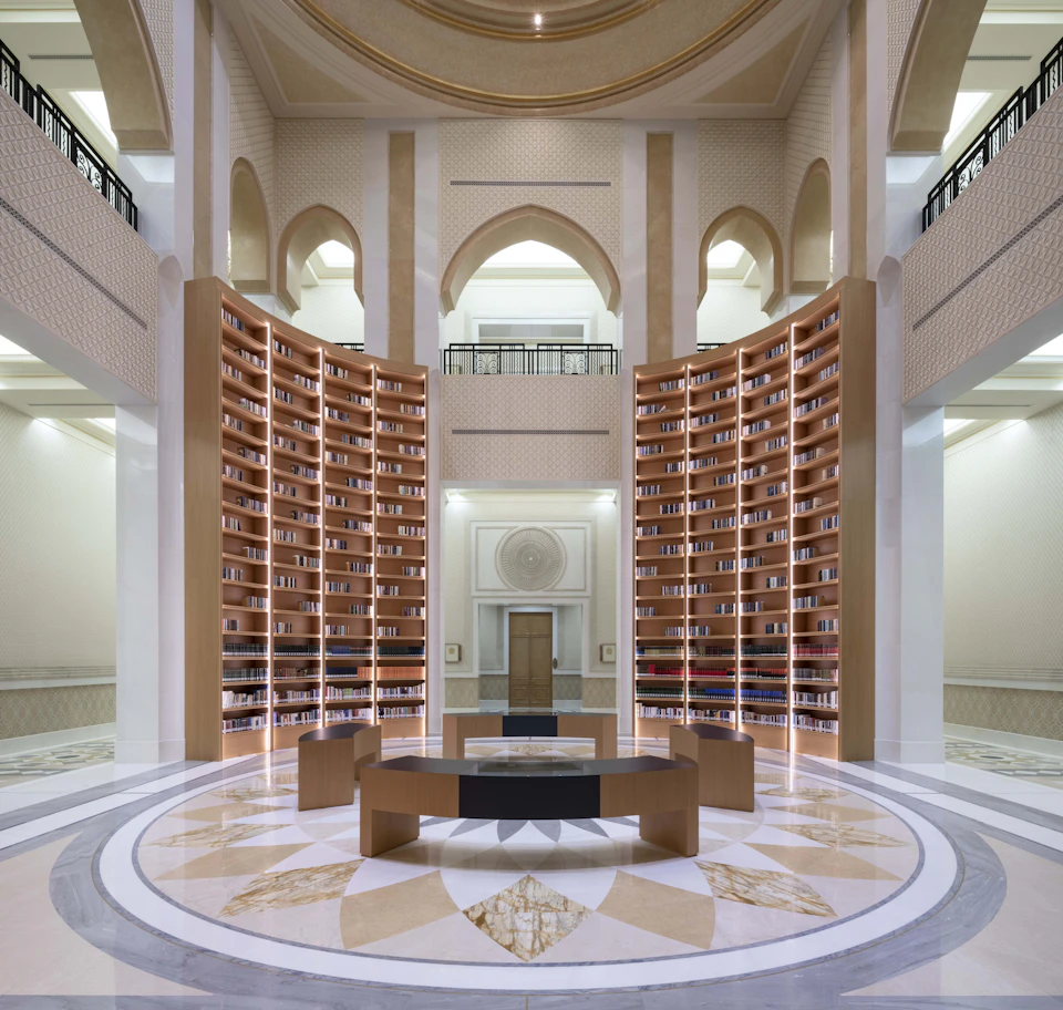 A grand library interior with high, arched ceilings, tall curved bookshelves, a circular desk, and ornate floor patterns.