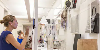 파란색 드레스를 입은 여성이 현대적인 갤러리의 디스플레이와 상호 작용하며, 선반에 다양한 물건이 있고 벽에 자전거가 장착되어 있습니다.