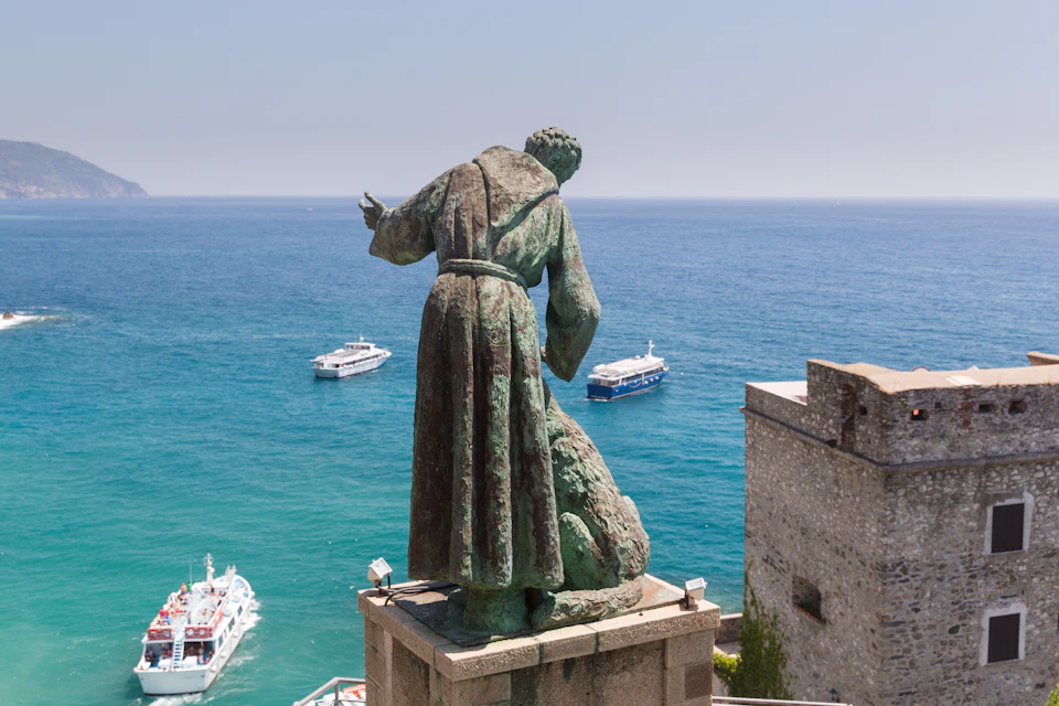Bronze statue of a person in robes overlooking a blue ocean with three boats, next to a stone structure.