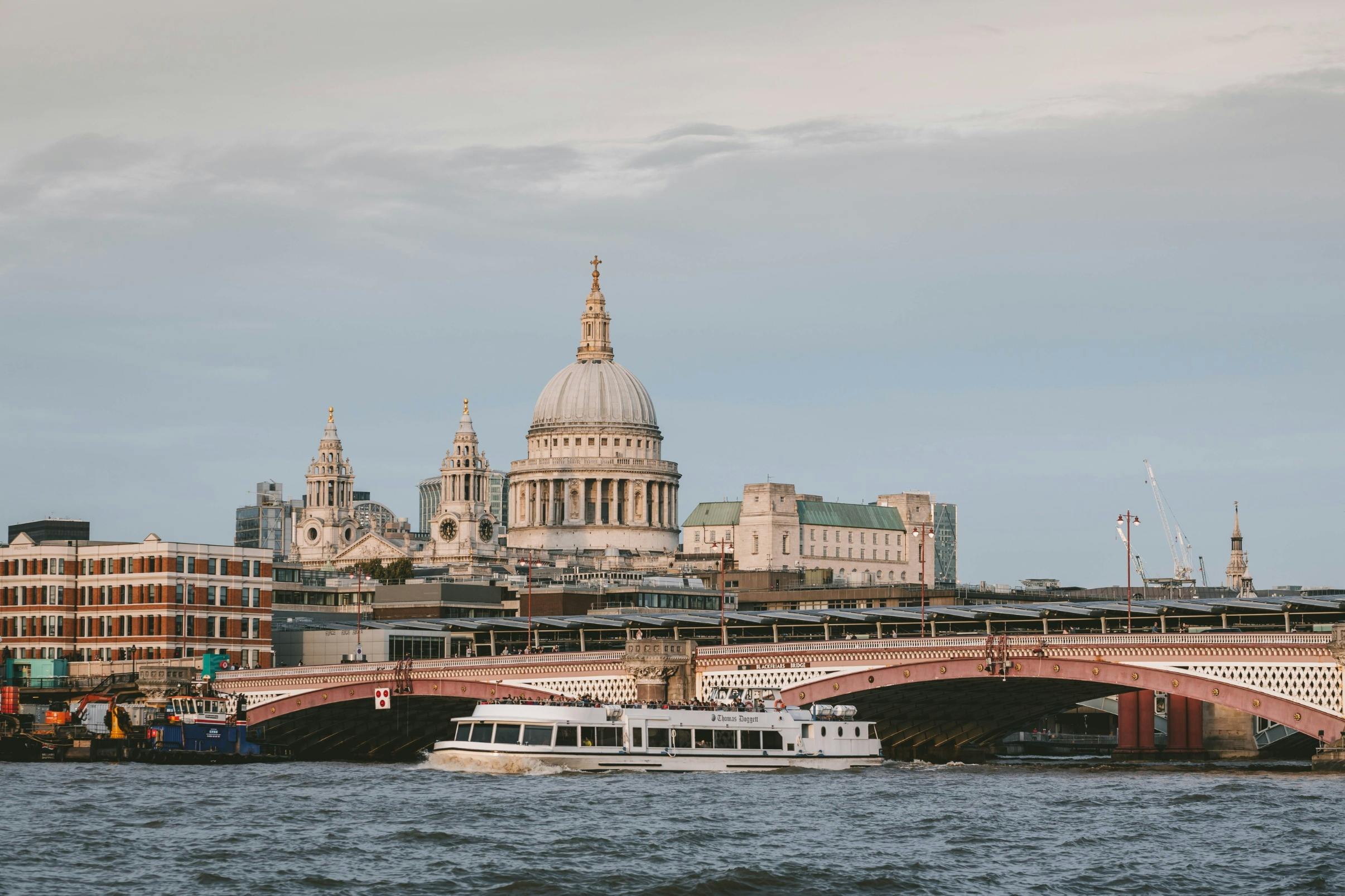 En flodbåt som passerar under en bro med St Paul's Cathedral och omgivande byggnader i bakgrunden.
