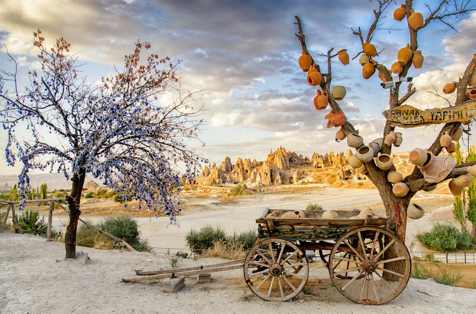 A rustic cart and a tree adorned with pots stand in a barren landscape with jagged rock formations in the background.