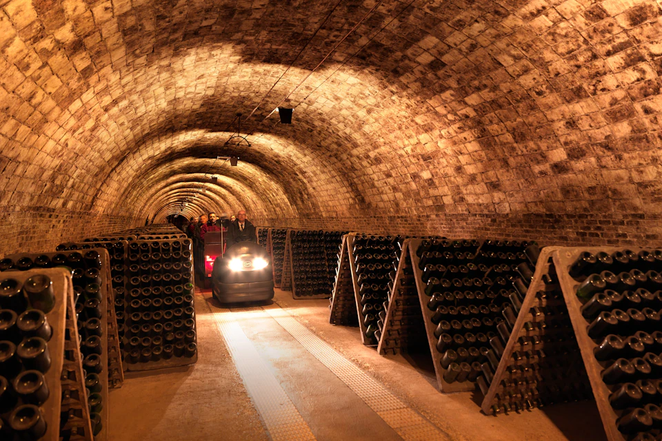 A dimly lit wine cellar with arched ceilings, stacks of wine bottles, and a small cart in the aisle.