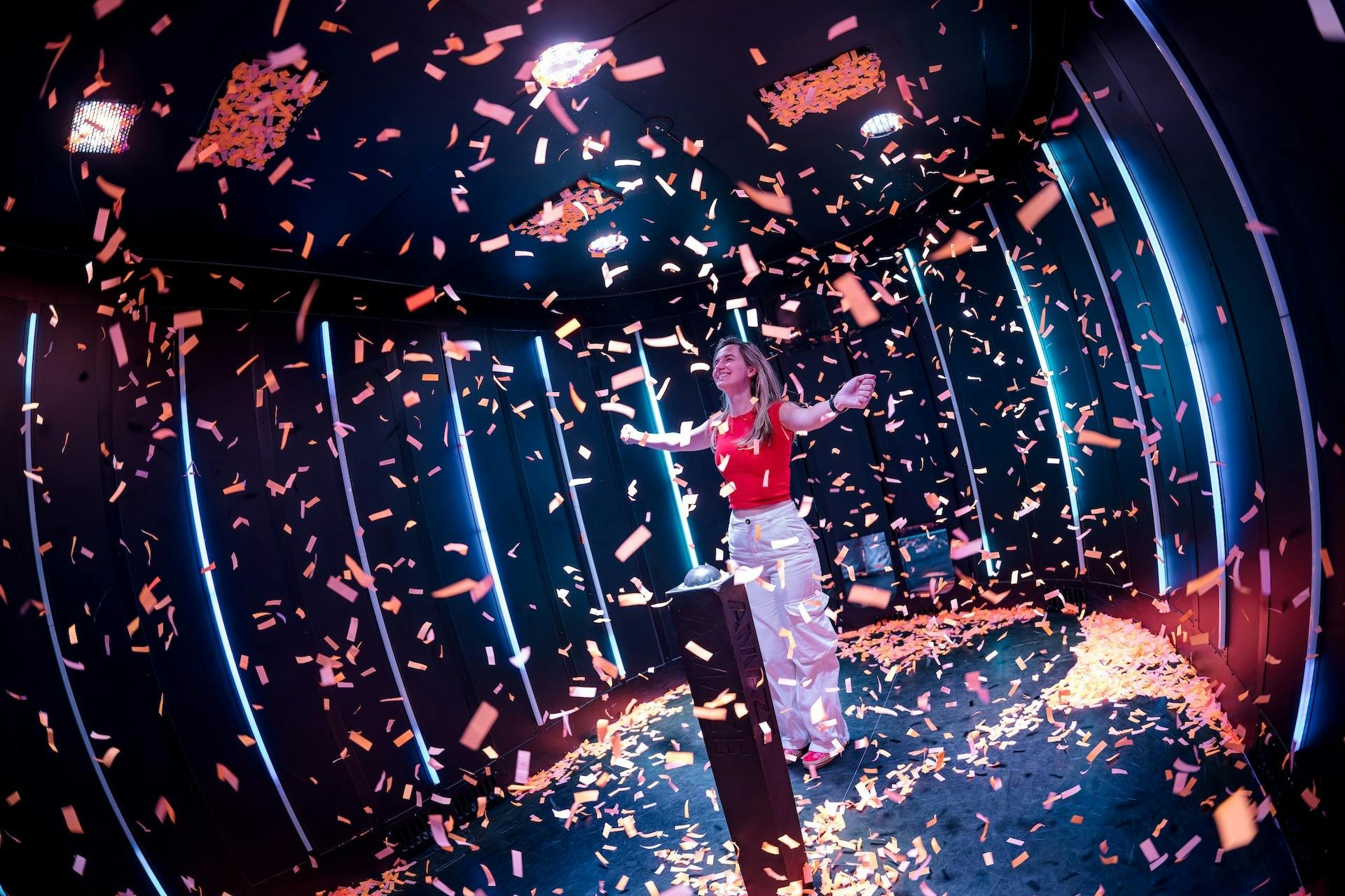 A person stands in a confetti-filled room with dark walls and vertical neon lights, arms raised in joy.